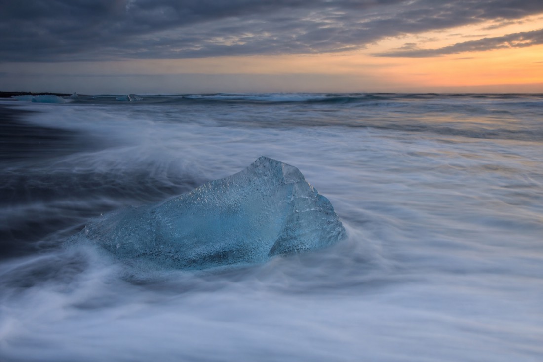 A long-exposure photograph of white surf blurring around a glowing block of glacier ice on the black sand of Diamond Beach at dusk