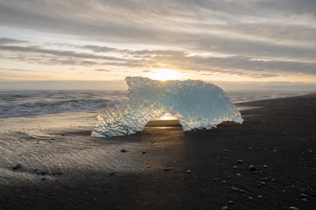 Low winter sun behind a pale-blue iceberg on black sand, the beach nearly empty of other ice