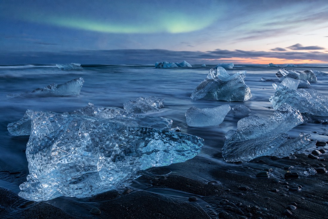 Blue-hour twilight over Diamond Beach Iceland with ice glowing on black sand under a faint aurora