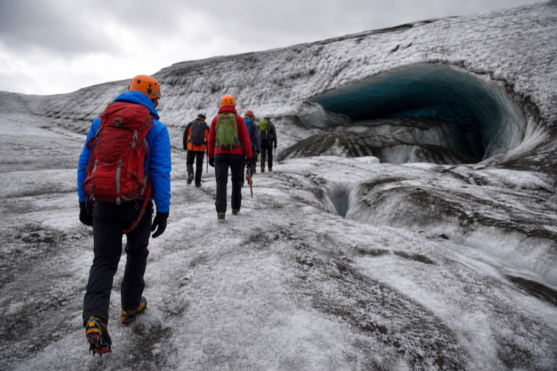 Certified glacier guide in crampons leading visitors across the snowy surface of Vatnajökull toward a natural ice cave entrance